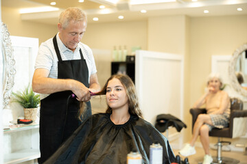 Elderly man, professional experienced hairdresser using curling irons to create curls and add volume to hair of young interested smiling girl..