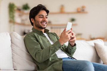 A young Arab man with a beard sits on a white couch while holding and looking at a white smartphone. He is wearing a green shirt and blue jeans.