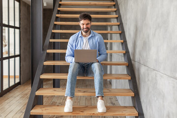 Poster - A man wearing a blue shirt and jeans sits on the wooden stairs in a modern home. He is working on a laptop computer, smiling. The stairway has a metal railing and leads to an upper level of the home.