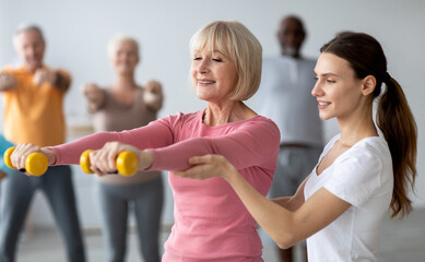 Wall Mural - Young lady coach helping senior woman while exercising with dumbbells, sporty multiracial group of elderly people having fitness class at nursing home, training with instructor