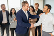 © Prostock-studio - A group of diverse business professionals stand together in a modern office setting. A smiling man in a blue suit extends his hand to shake the hand of a new colleague, who also smiles warmly