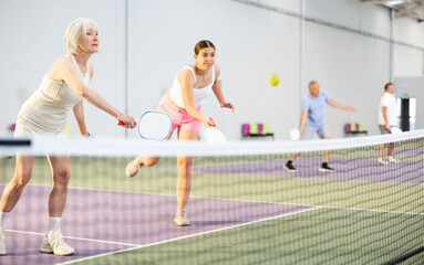 Wall Mural - Focused elderly woman playing friendly pickleball match in doubles team with young girl on indoors court. Sport and active lifestyle concept ..