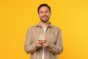 Poster - A man wearing a beige shirt stands against a bright yellow background, smiling as he holds a smartphone in his hands.