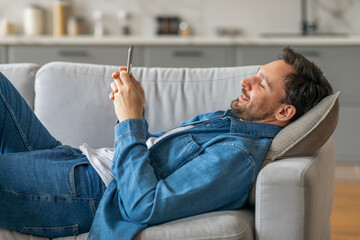 Poster - A man lies on a grey couch, propped up on a pillow, with his legs outstretched and his arms resting on the couch. He is using his smartphone with a smile on his face.