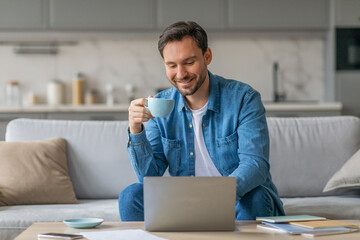 Poster - A man sits on a couch at home, working on a laptop while sipping a cup of coffee. He is wearing a casual blue shirt and smiling. He is working in a relaxed, comfortable setting.