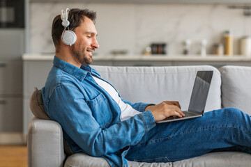 Aufkleber - A man in a denim shirt sits on a gray couch with his legs stretched out. He is wearing white headphones and looking down at his laptop while typing.