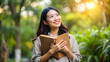 © Sniezka - Smiling Asian-looking woman holding a book in a lush garden during sunset. Suitable for educational content, wellness blogs, or nature-themed promotions. Warm and inviting color palette.