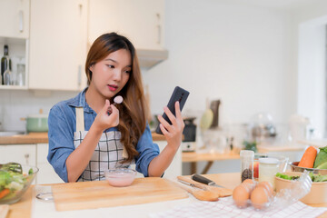 Wall Mural - Young asian woman eating yogurt and surfing social media on smartphone while preparing fresh vegetables salad and cooking healthy breakfast food in modern kitchen with healthy lifestyle at home