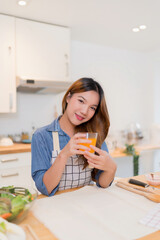 Wall Mural - Young asian woman holding glass of orange juice and drinking fresh fruits while preparing vegetables salad and cooking healthy breakfast food in modern kitchen with healthy lifestyle at home