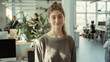 © VK Studio - A woman stands in a bright, modern office, smiling warmly at the camera. The airy space is filled with plants and colleagues working in the background.