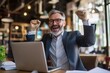 © Антон Сальников - An older business man wearing a suit celebrates success in his office holding papers and looking at his laptop, exulting over the company's growth and success.