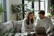 © dakinya - Happy couple working on laptop, bright living room, casual attire, green plants, natural light, cozy home environment, copy space