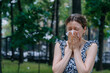 © wifesun - Woman sneezes into a tissue while standing outside in a park, surrounded by airborne allergens like poplar fluff.