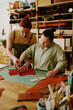 © AnnaStills - Highly skilled male tailor helping his new female intern with down syndrome to cut out leather workpiece
