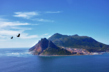 Wall Mural - Blue sky, clouds and mountain or ocean with buildings in nature or fishing village by sea. Coastal town, birds and waves or landscape in summer for travel, vacation and outdoor for holiday by beach