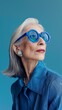 © Maria Mikhaylichenko - Elegant senior woman with silver hair wearing vibrant blue sunglasses and jacket, exuding confidence against a teal background. Fashion portrait of ageless style.