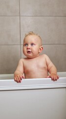 Wall Mural - A cute blue-eyed 1-year-old boy is bathing in a white bath. Baby's personal hygiene. A child with a smile on his face while bathing. Vertical video