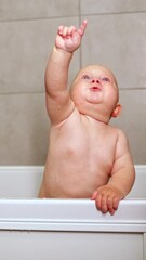 Wall Mural - A cute blue-eyed 1-year-old boy is bathing in a white bath. Baby's personal hygiene. A child with a smile on his face while bathing. Vertical video