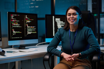 Portrait of smiling young woman wearing glasses as female computer programmer sitting at office workplace and looking at camera copy space