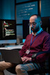 © Mediaphotos - Vertical portrait of adult man using laptop sitting at office workplace in cybersecurity department with code lines and blue neon light