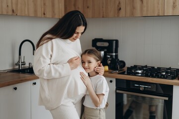 a pregnant woman and her 6-year-old daughter hug and smile happily in their kitchen, the daughter strokes her mother's tummy with her hand