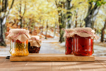 Wall Mural - Jars of autumn preserves of vegetables and fruits on an autumn table with an empty space for products. In the background, an autumn garden and trees with yellow and orange leaves