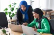 © Studio Marmellata - Two women in hijabs collaborating on a project in a modern office, using laptops and sticky notes to outline their strategies.
