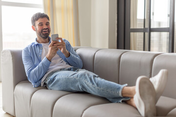 Poster - A man in a blue shirt and jeans is sitting on a gray couch. He is relaxing, looking at his phone and smiling. His feet are up on the couch and he is wearing white sneakers.