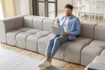 Poster - A man sits on a modern gray sectional sofa in a living room. He is looking at a smartphone and smiling, while using a laptop. He is wearing jeans and a blue button-up shirt, high angle view