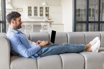Poster - A man wearing a light blue shirt and jeans is lying on a grey couch. He is working on a laptop, while his feet are propped up on the couch in front of him. He appears to be relaxed