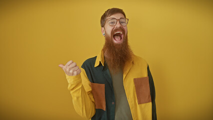 Wall Mural - A cheerful young bearded man with red hair wearing glasses gestures thumbs-up against a vibrant yellow background.