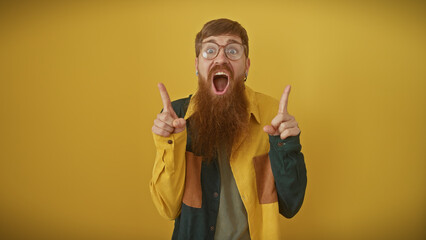 Wall Mural - Excited bearded redhead man pointing upwards with a shocked expression isolated against a yellow background.