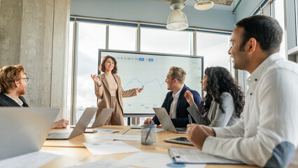 Aufkleber - A businesswoman, standing in front of a whiteboard with a graph displayed, gestures towards the data as she presents information to a group of colleagues seated at a table in a modern office