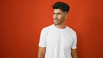 Handsome young hispanic man with beard in casual white t-shirt against an isolated red wall background.