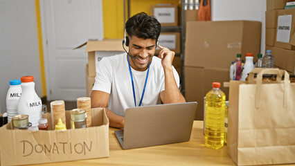 A cheerful man in a warehouse organizes donations while working on a laptop and wearing a headset