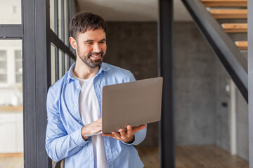 Poster - A man stands in a modern office space, smiling as he uses his laptop. He is dressed in a casual blue shirt and is leaning against a glass wall. He has short, dark hair and a beard.
