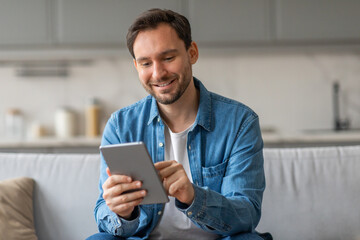 Poster - A man, wearing a blue denim shirt, sits on a white couch in a living room setting. He holds a tablet in his hands and scrolls through it with a smile on his face.