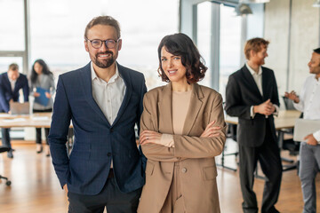 Aufkleber - A man and woman in business attire stand confidently in front of a large window. They are both smiling and appear to be happy and successful. The background is blurred and features other people