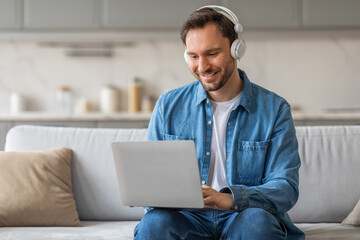 Poster - A man in a denim shirt sits on a couch in a living room. He is using a laptop computer and smiling. He is wearing headphones and is focused on his work.