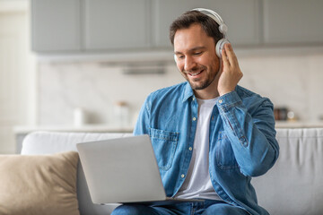 Poster - A man wearing headphones sits on a couch in a living room while smiling and using a laptop. He is wearing a blue denim shirt over a white t-shirt and blue jeans.