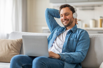Poster - A man is sitting on a light grey couch, leaning back with one hand behind his head, wearing headphones and smiling as he uses a laptop.