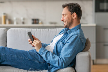 Poster - A man is sitting on a couch in a living room while using a smartphone. He is smiling as he looks at the phone. He is wearing jeans and a blue denim shirt.