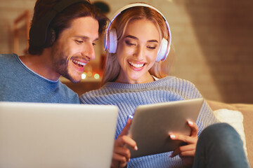 Wall Mural - Happy Man And Woman Using Digital Tablet Computers Wearing Headphones Sitting On Sofa Indoor In Evening