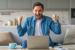 © Prostock-studio - A man wearing a blue shirt and jeans is sitting on a sofa, looking at a laptop with a wide smile and both fists raised in the air. He seems to be celebrating a success or achievement