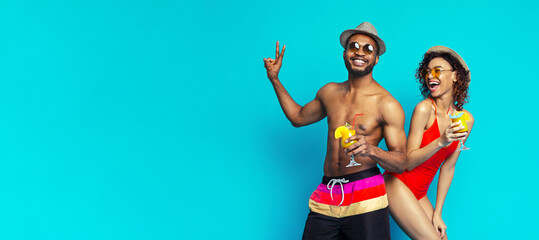 Wall Mural - A happy black couple pose with cocktails against a bright blue backdrop. The man is shirtless, wearing colorful swim trunks, and a hat. The woman is wearing a red swimsuit and a straw hat