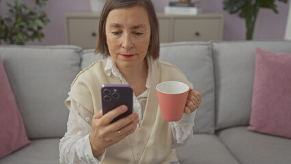 Sticker - Mature woman holding mug while using smartphone on sofa indoors