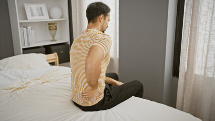 Wall Mural - A young hispanic man with a beard experiences back pain while sitting on a bed in a well-lit bedroom.