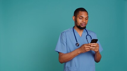 Male nurse using mobile phone to scroll on social media feed during hospital job shift break. Healthcare worker using smartphone to text friends, studio backdrop, camera B