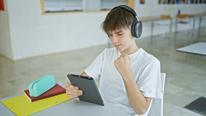 Wall Mural - Blond teenager male sitting at a table in a library, studying on a tablet with headphones and notebooks.