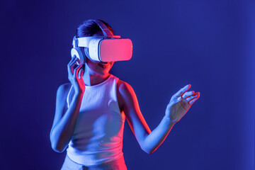 Smart female standing with surrounded by cyberpunk neon light wear VR headset connecting metaverse, futuristic cyberspace community technology. Woman using hand touching virtual object. Hallucination.